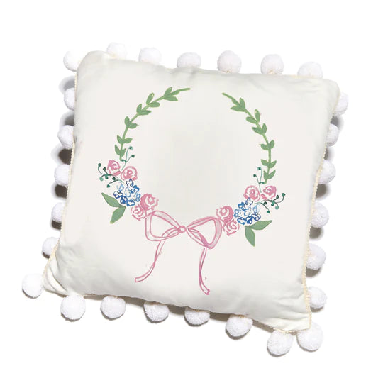 A square pillow with a white background featuring a floral monogram design in colors green, blue, and pink, surrounded by a border of white pompoms.
